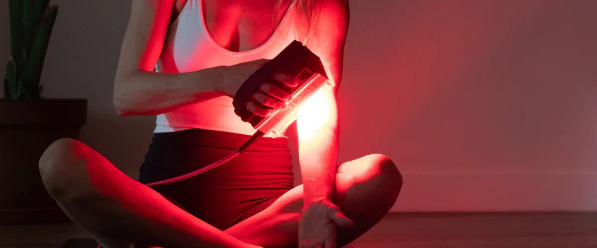Woman sitting holding a Chroma Trinity light therapy device close to her forearm emitting a red glow against a dark grey background