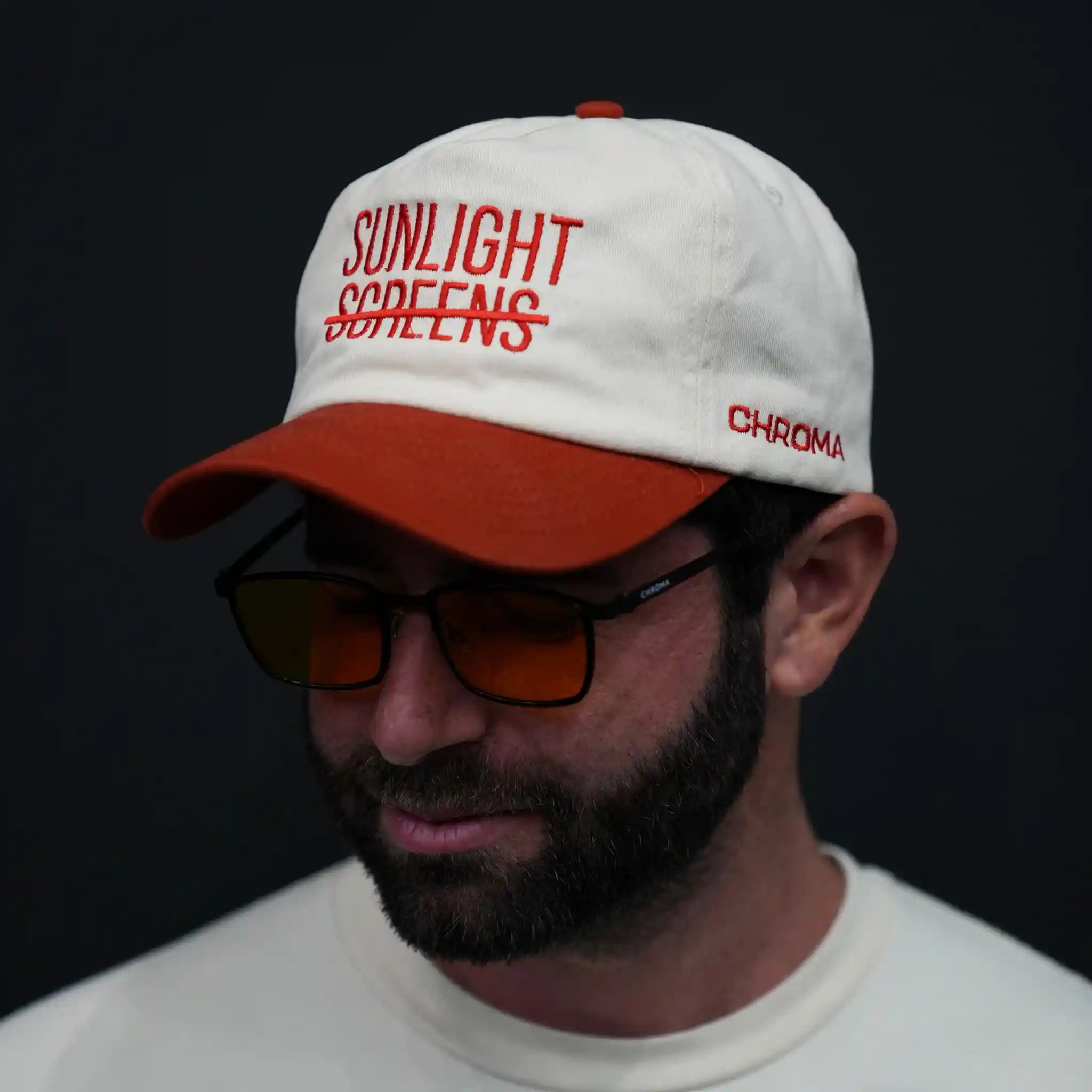 Man wearing a white cap with red text (sunlight screens) and (Chroma) branding on the side, also wearing Chroma nightshades glasses against a black background