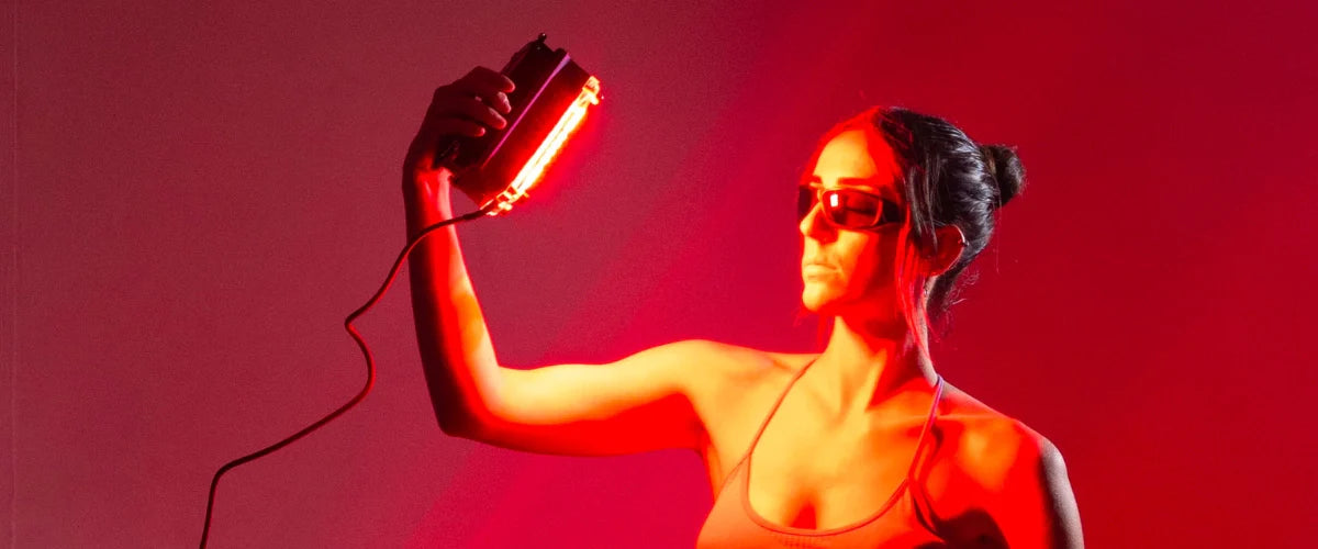 Woman holding a Chroma D-Light light therapy device towards her face at a high angle emitting a red light against a dark red background