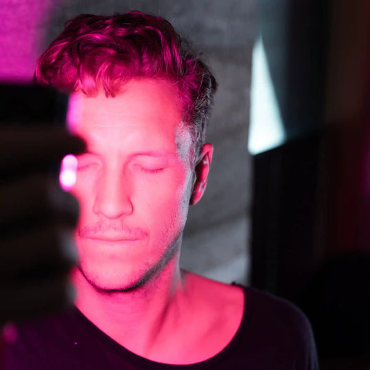 Man holding a Chroma Radiance light therapy device towards his face emitting a pink hue against a grey background.