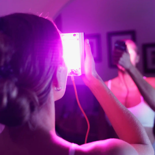 A woman holding the Chroma Radiance light therapy device towards her face emitting a pink hue. She is facing away from the camera towards a mirror