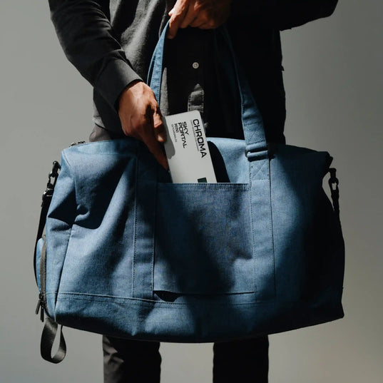 Person holding a blue duffel bag and taking out a Chroma Sky Portal Mini out of the bag with a gray background