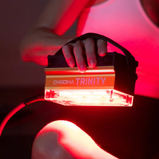 Hand holding a light therapy device labeled 'Chroma Trinity' with red lighting in the background