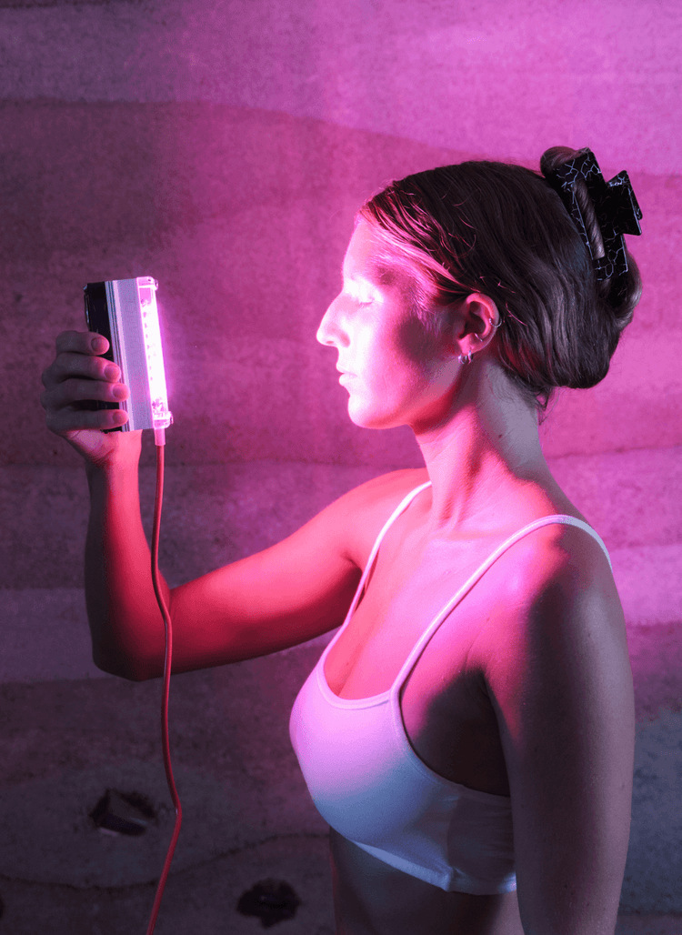 Woman holding a Chroma Radiance light therapy device emitting pink light against a textured wall.